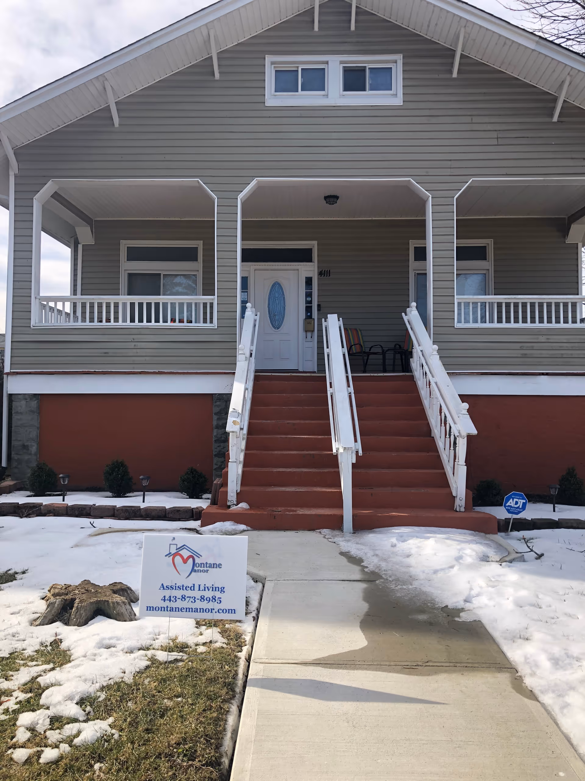 Front exterior view of Montane Manor Assisted Living building with a gray facade, white trim, and a central staircase leading up to a white door. There is a sign on the lawn with the facility's name, phone number, and website. Snow is partially covering the ground around the walkway.
