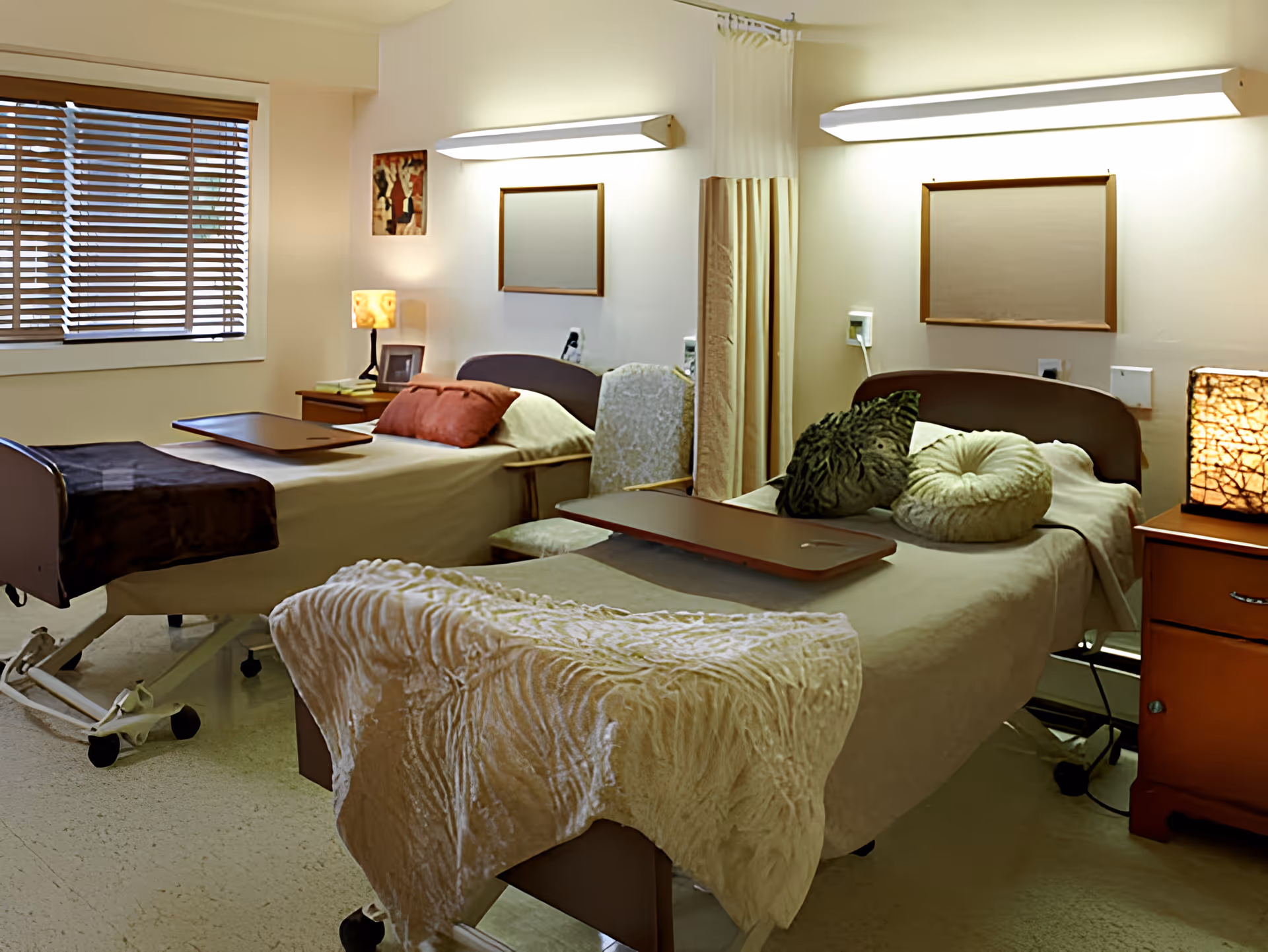 A shared bedroom in a healthcare facility with two hospital-style beds, each with pillows and blankets. There are bedside tables with lamps, a window with blinds, and a privacy curtain between the beds. The room has soft lighting and a calm atmosphere.