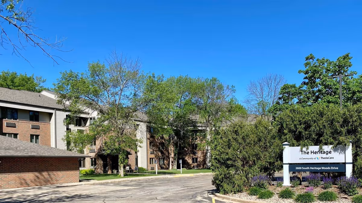 Exterior view of The Heritage senior living facility on a clear sunny day, showing a multi-story brick and siding building surrounded by trees and greenery, with a sign in front displaying the facility name and address.