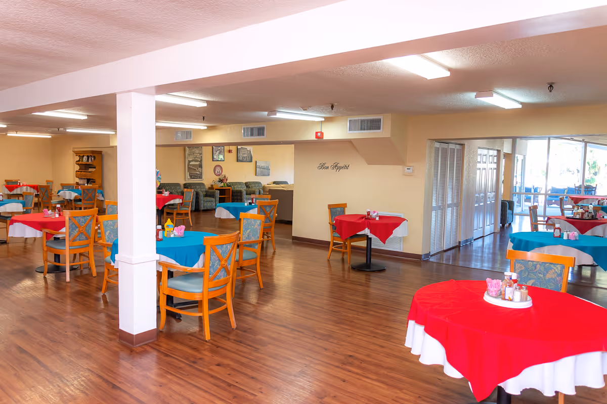 A spacious dining room with wooden floors and multiple round tables covered with red and blue tablecloths. Each table has wooden chairs with patterned cushions and a centerpiece with condiments. The room has beige walls, fluorescent ceiling lights, and a seating area with green patterned sofas in the background. A wall decal reads 'Bon Appétit'.