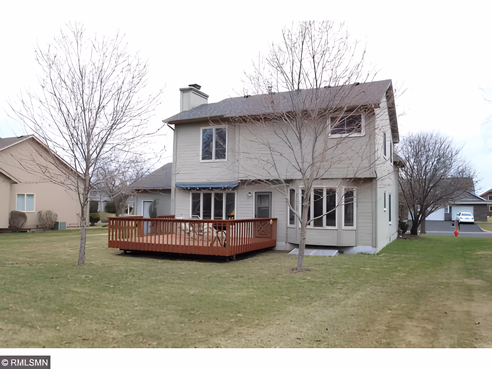 Rear exterior view of a two-story house with a wooden deck, leafless trees, and a grassy yard in a residential neighborhood.