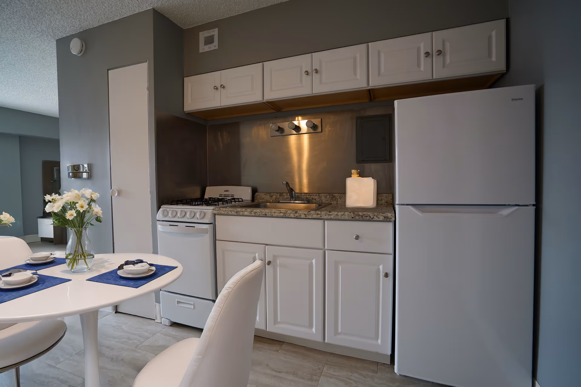 A compact kitchen area with white cabinets, a small white gas stove, a stainless steel sink, and a white refrigerator. A round white dining table with white chairs is partially visible in the foreground, set with blue placemats, white bowls, and a vase of white flowers. The walls are painted gray and the floor has light-colored tiles.