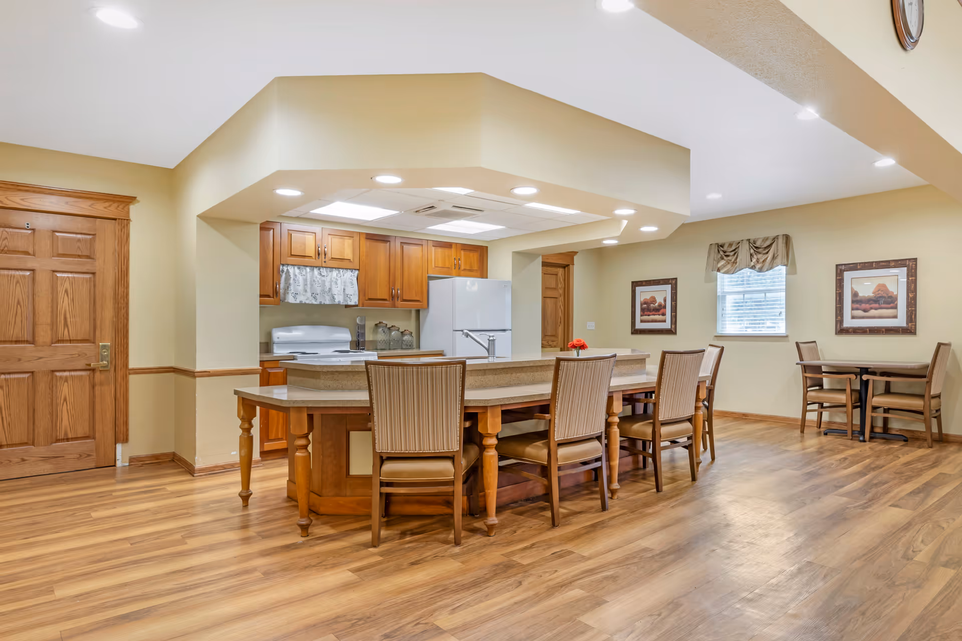 A spacious kitchen and dining area in a senior living facility with wooden flooring and beige walls. The kitchen features wooden cabinets, a white refrigerator, and a white stove. A large island with a beige countertop has several chairs around it. There are two framed pictures on the wall and a window with a valance letting in natural light. A small dining table with chairs is visible in the background.