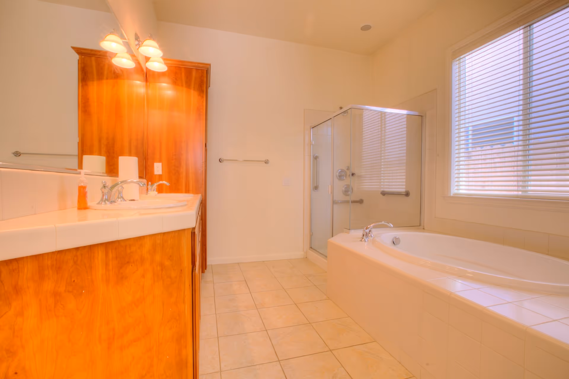 A clean and spacious bathroom featuring a large bathtub with tiled surround, a glass-enclosed shower, a wooden vanity with a sink, a mirror, and wall-mounted lights above the mirror. There is a window with blinds allowing natural light into the room.