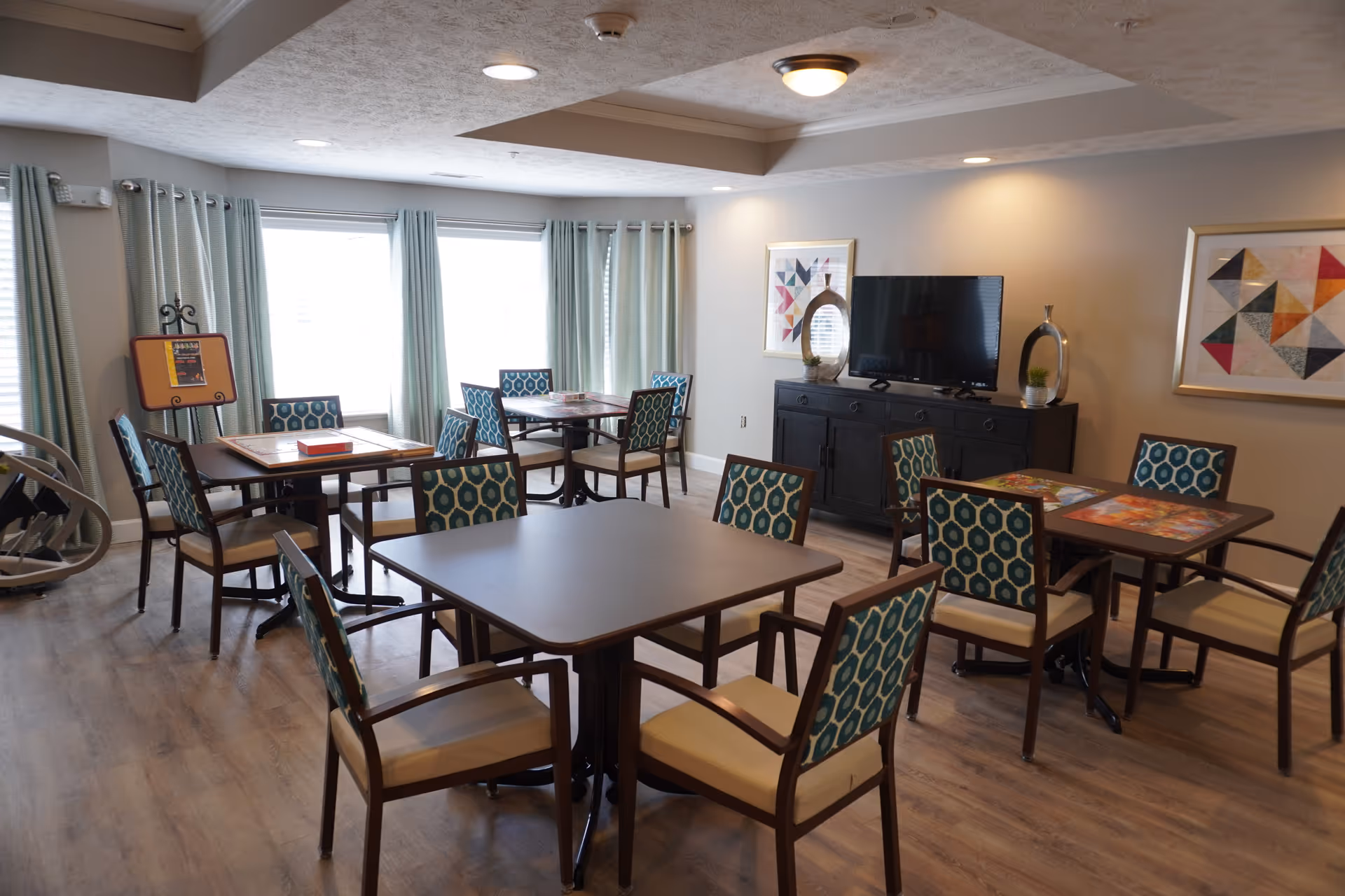 A well-lit common room with multiple square tables and chairs arranged for group seating. The chairs have patterned blue and beige upholstery. Large windows with light curtains allow natural light to fill the room. A black cabinet against the wall holds a flat-screen TV and decorative items, with two framed abstract artworks hanging above it. The room has a light wood floor and a ceiling with recessed lighting.