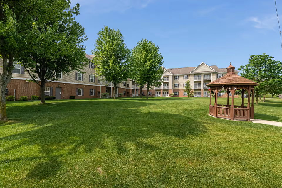 A large green lawn with several trees and a wooden gazebo on the right side, in front of a multi-story residential building under a clear blue sky.