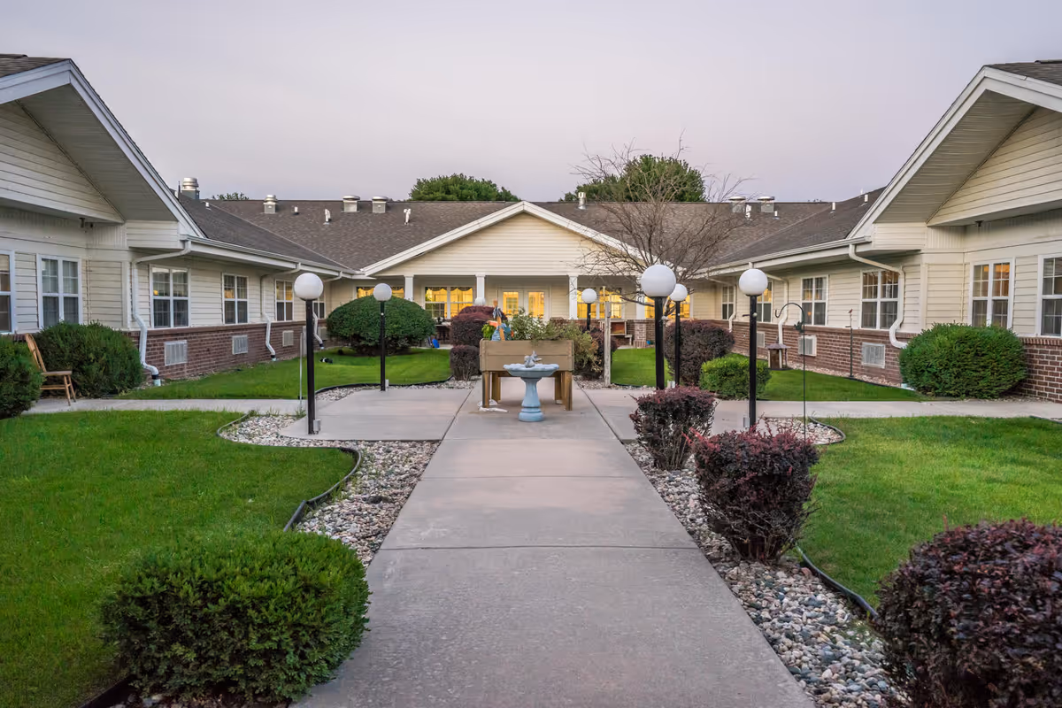 Outdoor courtyard area of Saunders House featuring a concrete walkway lined with round globe lamp posts, green grass, bushes, and a central water fountain with a bench. The building surrounds the courtyard with beige siding and brick accents under a cloudy sky.
