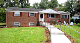 Single-story brick house with a white front door and a concrete ramp with metal railings leading up to the entrance. The house has several windows with white shutters and is surrounded by a green lawn with trees in the background. Two white chairs are placed on the grass to the right of the ramp.