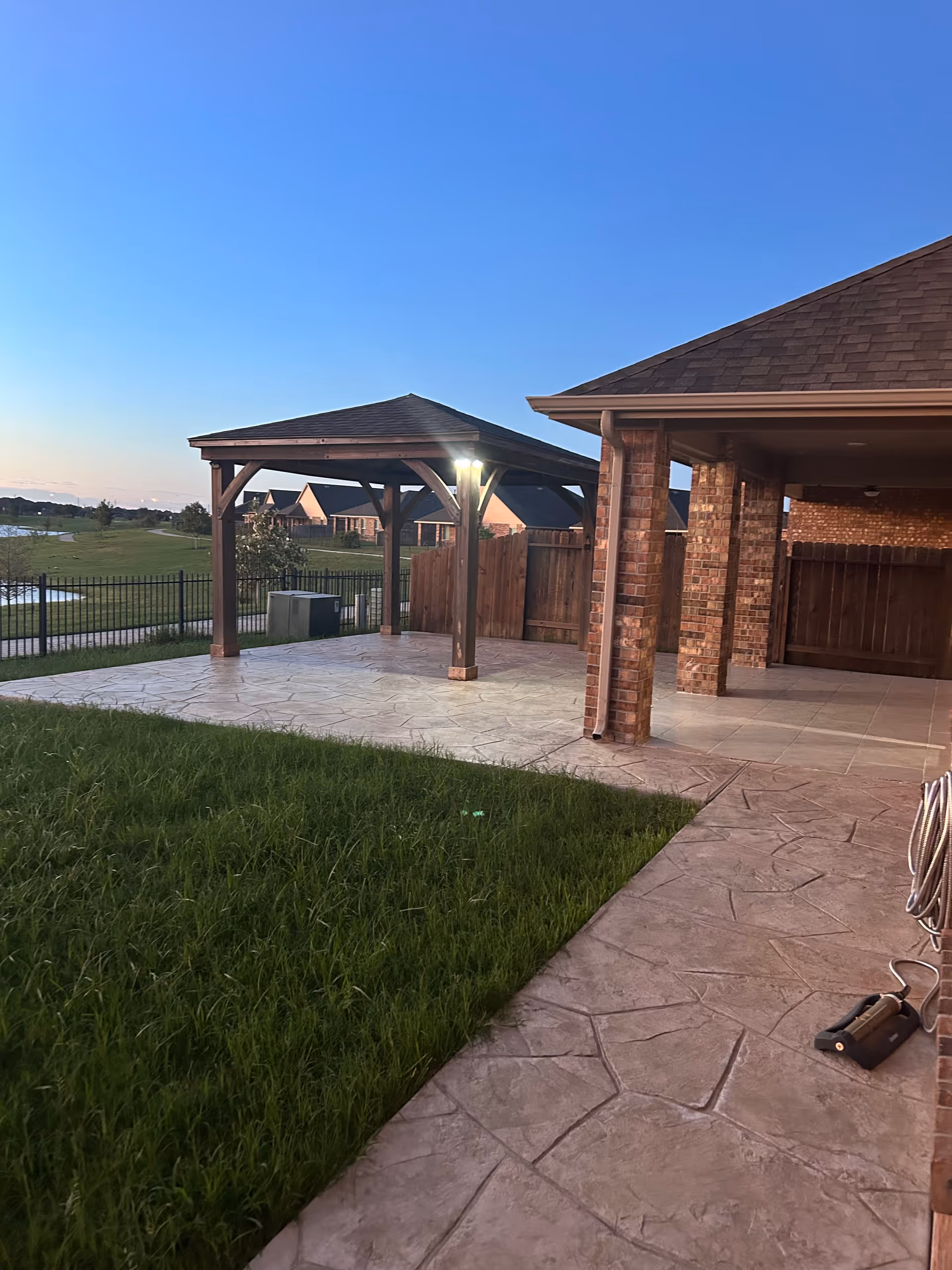 Outdoor patio area with a wooden gazebo structure and a covered brick porch. There is a grassy lawn adjacent to the paved patio, and a black metal fence enclosing the area. The sky is clear with a hint of sunset light.