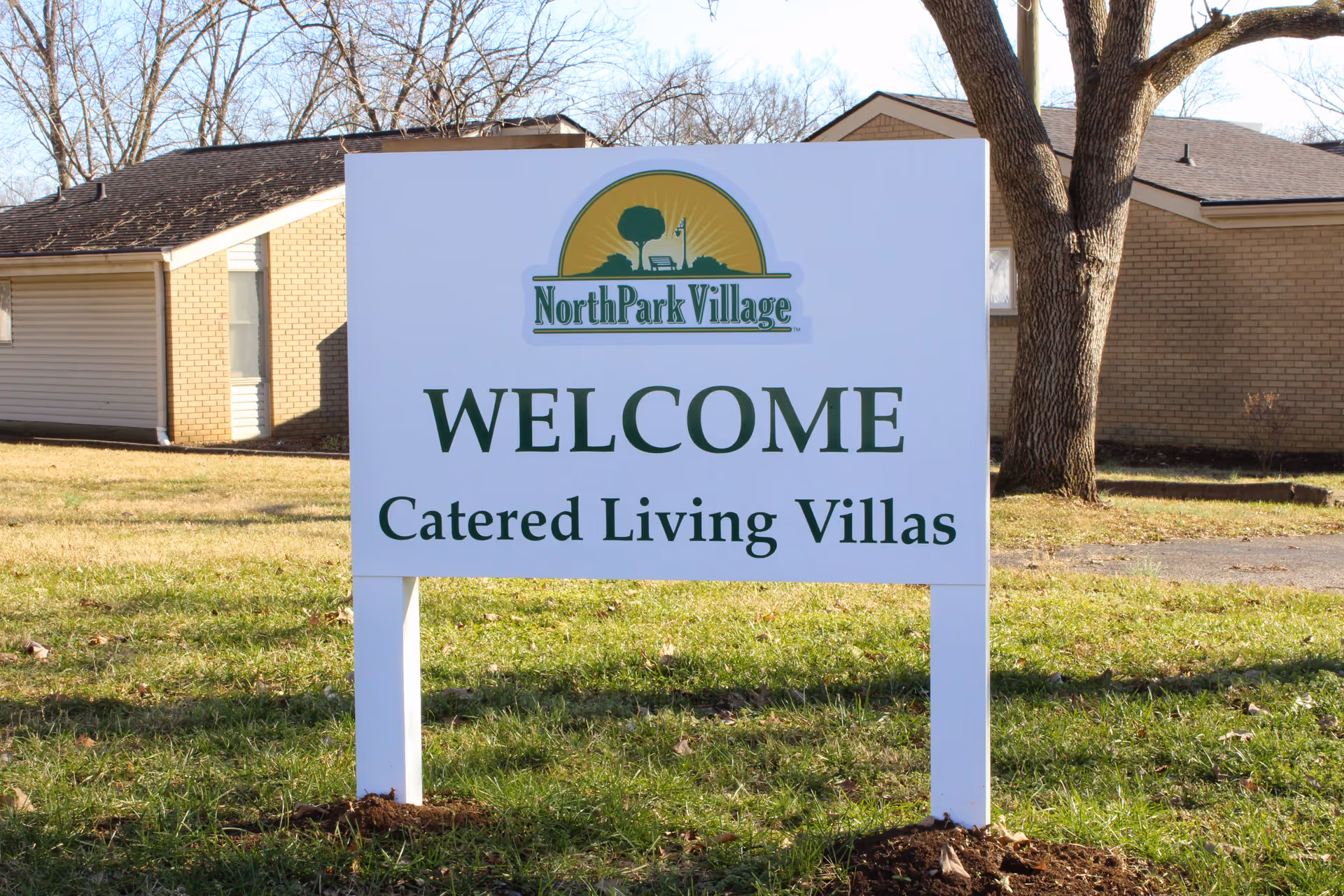 A white sign on a grassy area in front of a building reads 'North Park Village WELCOME Catered Living Villas' with a logo featuring a tree, bench, and streetlamp above the text.