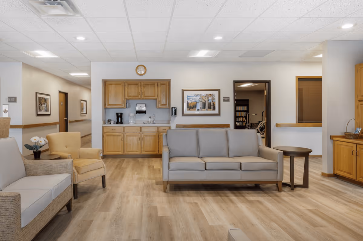 A senior living facility common area with light wood flooring, beige walls, and a drop ceiling with recessed lighting. The room features a gray three-seat sofa, a beige armchair, a small round wooden side table, and a wicker loveseat with light cushions. On the back wall, there is a wooden cabinet with a coffee maker, water dispenser, and a clock above. A framed picture hangs on the wall next to an open doorway leading to another room with exercise equipment. There is also a hallway with framed pictures on the left side.