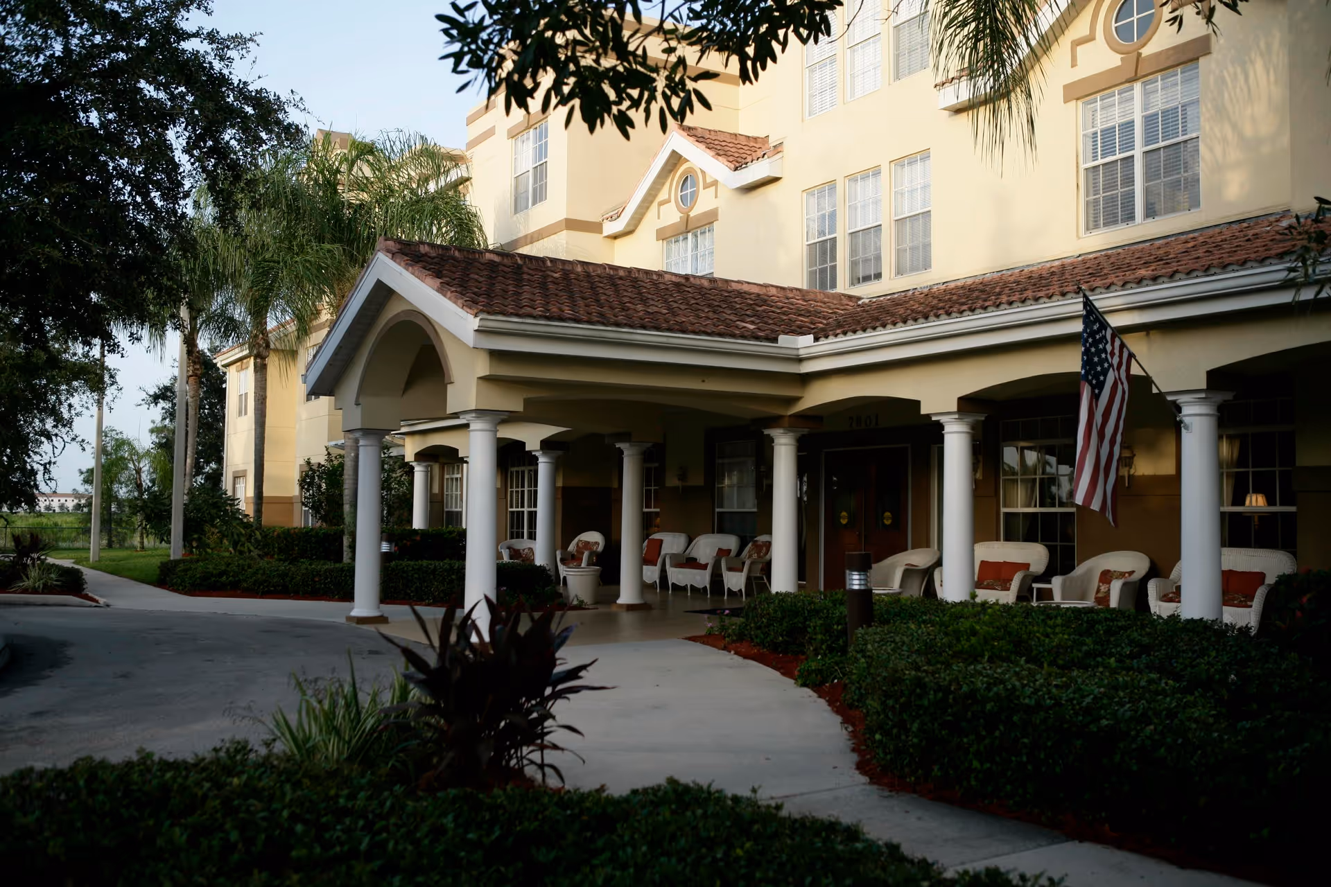 Exterior view of a senior living facility with a covered entrance supported by white columns. Several white wicker chairs with red cushions are arranged on the porch. An American flag is displayed near the entrance. The building is surrounded by well-maintained bushes and palm trees.