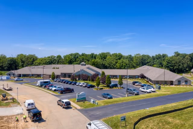 Aerial view of Cedarhurst Senior Living of Waterloo, showing a large single-story building with a parking lot filled with cars. The building is surrounded by green grass and trees in the background under a clear blue sky.