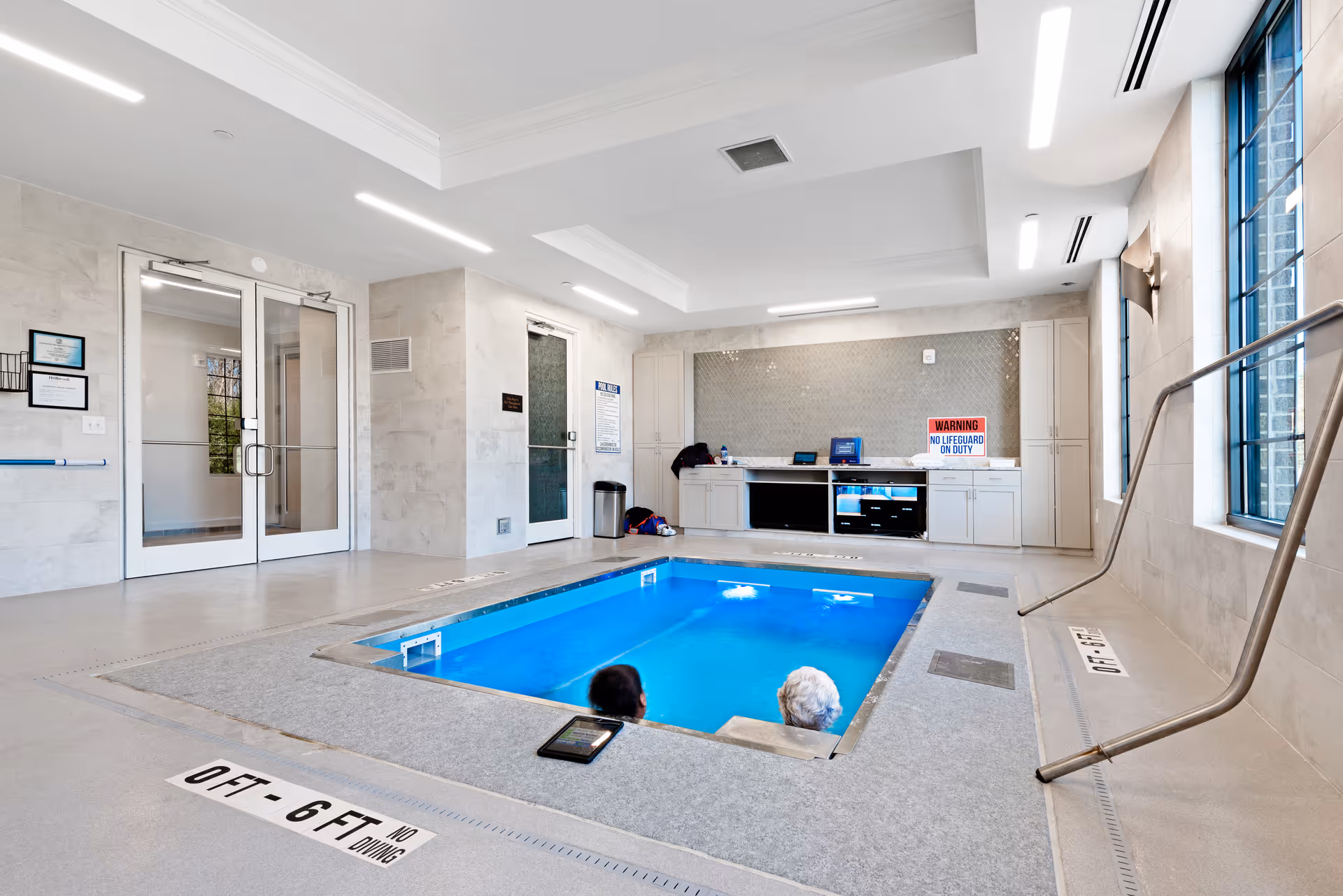 Indoor pool area with two people partially submerged in the water. The room has light-colored tiled walls and floors, large windows on the right side, and a built-in cabinet with a countertop and various items including a warning sign that says 'No Lifeguard On Duty'. There are double glass doors on the left side and a metal handrail leading into the pool on the right.