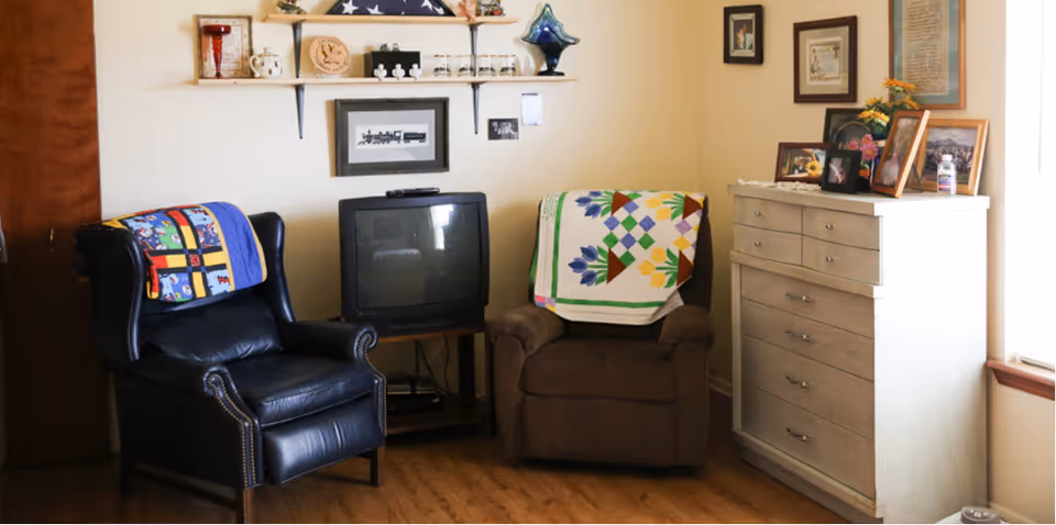 A cozy living room corner with two armchairs, one black leather and one brown fabric, each draped with colorful quilts. Between the chairs is a small wooden TV stand holding an older-style television. Above the TV are two wall shelves displaying various decorative items, including a folded American flag, a clock, and small figurines. To the right is a white chest of drawers topped with framed photos and other decorations. The room has wooden flooring and light-colored walls with framed pictures hanging.