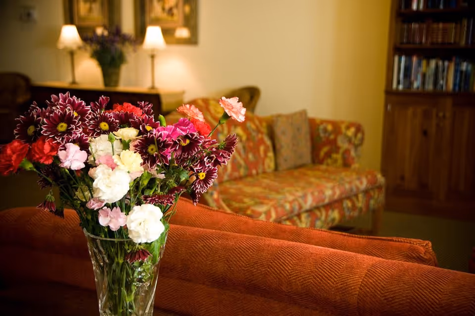 A vase of mixed flowers on a table in front of a patterned sofa in a cozy living room with lamps and a bookshelf.