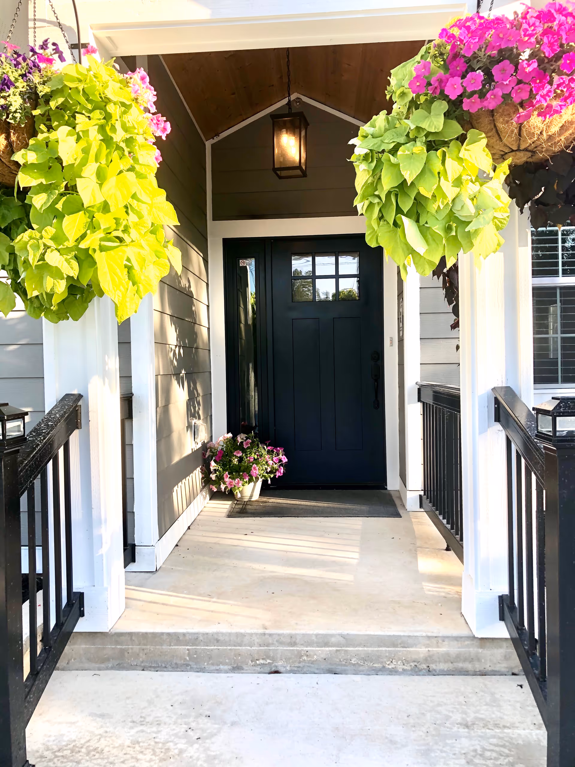 Front porch entrance with a dark blue door, hanging flower baskets, a potted plant, and black railings.