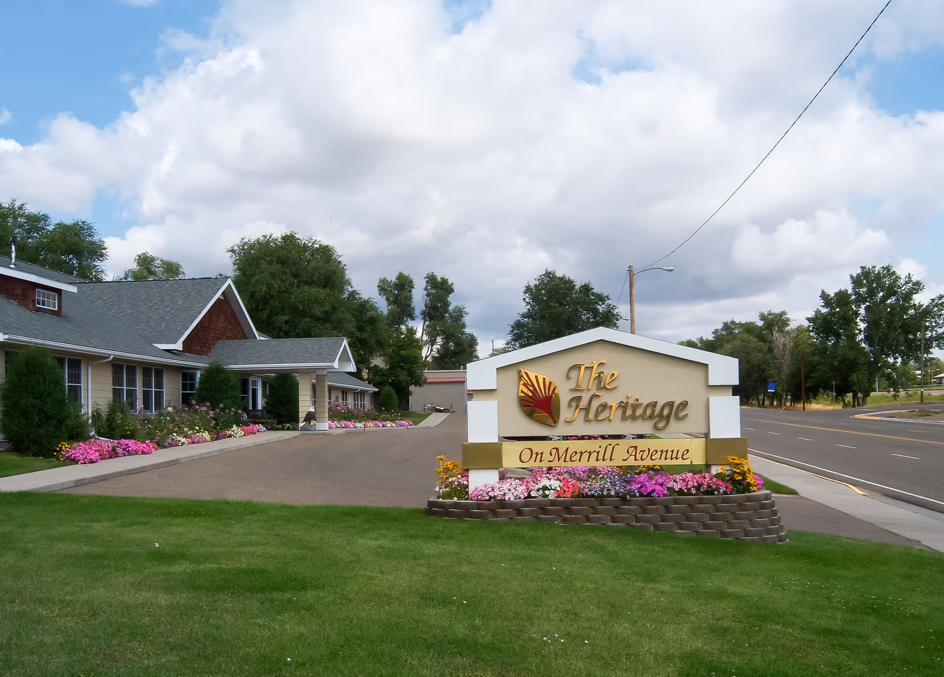 Exterior view of The Heritage senior living facility on Merrill Avenue, showing a large sign with the facility's name surrounded by colorful flowers and a driveway leading to the building with a well-maintained lawn and trees in the background under a partly cloudy sky.