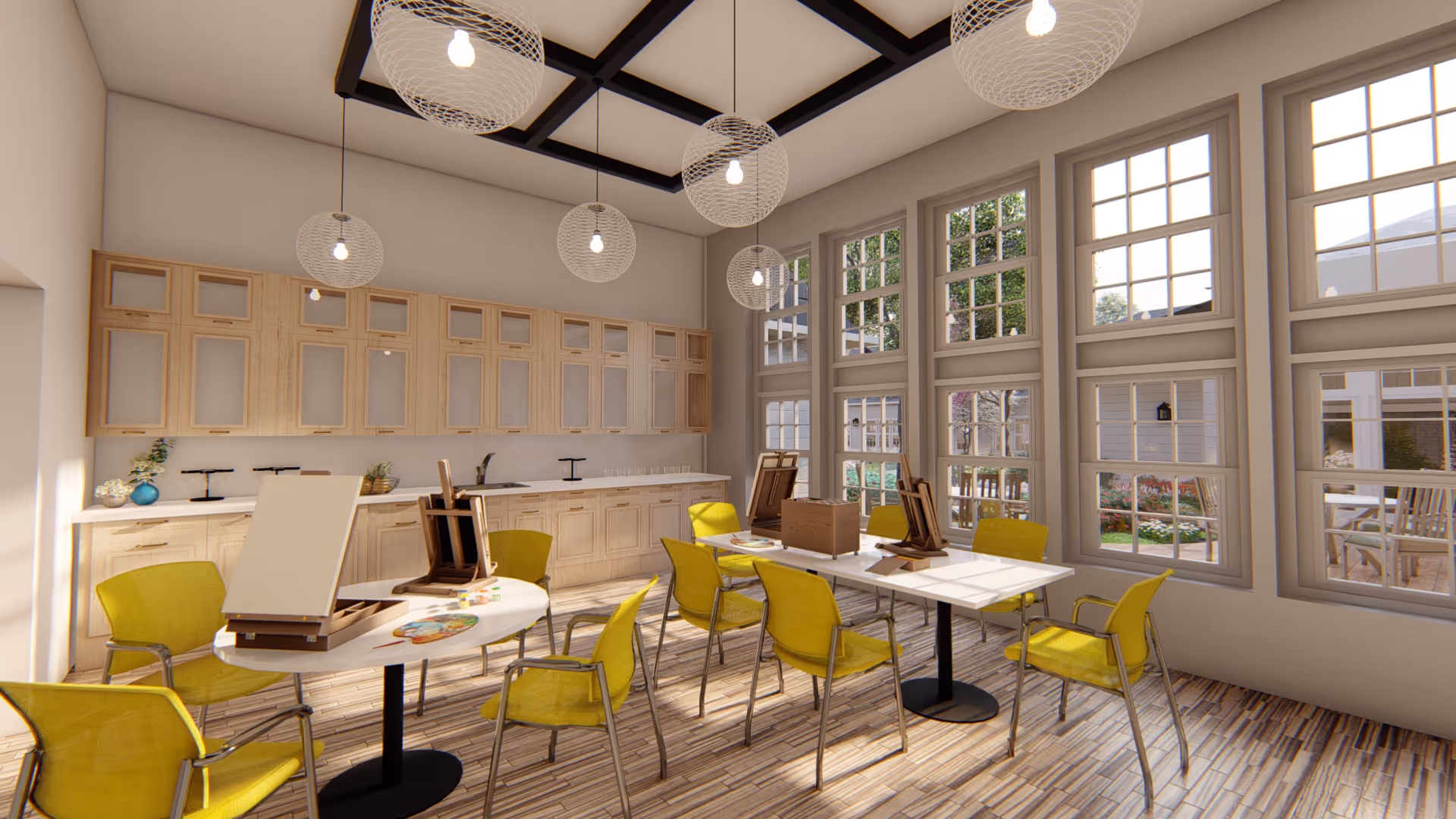 A bright and spacious activity room with large windows letting in natural light. The room features multiple tables with yellow chairs arranged around them. On the tables are easels and painting supplies, suggesting an art or craft space. The back wall has light wood cabinetry and a countertop with decorative items. Modern pendant lights hang from a ceiling with dark beams.