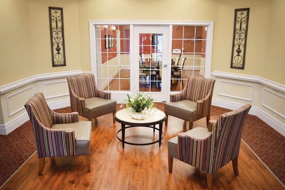 A cozy seating area with four striped armchairs arranged around a round coffee table with a floral centerpiece. The room has wooden flooring, beige walls with white wainscoting, and two decorative wall hangings. In the background, there is a glass door leading to a dining room with a table and chairs.