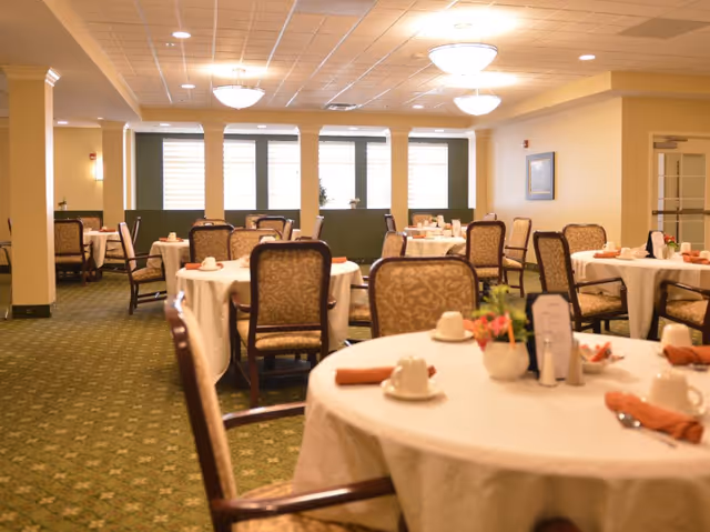 A bright dining room with round tables set with white tablecloths, chairs, and place settings.