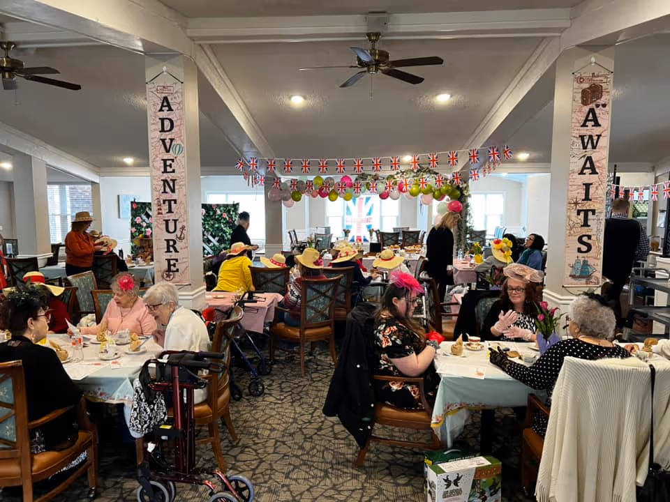 A lively dining room in The Pines Senior Living decorated with colorful balloons and British flags. Several elderly residents wearing festive hats are seated at tables enjoying tea and conversation. Two large vertical banners on pillars read 'ADVENTURE' and 'AWAITS'. The room is well-lit with ceiling fans and has a cozy, welcoming atmosphere.