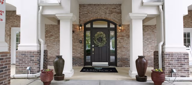 Entrance to a building with a black door decorated with a green wreath, flanked by two wall-mounted lantern lights. The entrance is framed by white columns and brick walls, with four large decorative pots containing plants placed symmetrically on the ground.