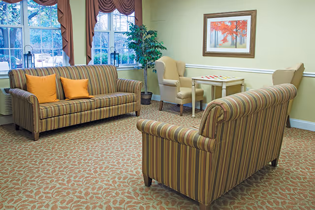 A cozy living room area with two striped sofas facing each other, two beige armchairs around a small table, a large window with brown curtains, a potted plant, and a framed picture of a tree with red leaves on a light green wall.