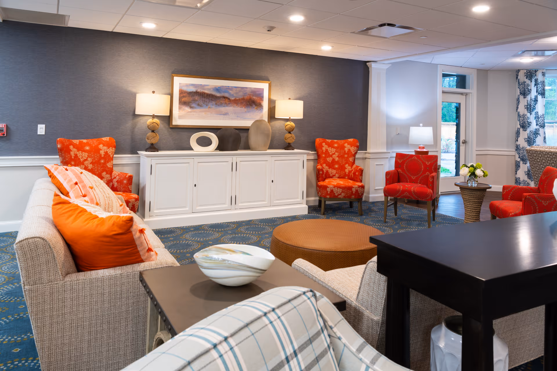 A cozy living room area with a beige sofa adorned with orange and striped pillows, two red patterned armchairs, a round brown ottoman, and a white cabinet against a gray textured wall. The cabinet holds decorative vases and is flanked by two table lamps. A framed landscape painting hangs above the cabinet. The room has blue patterned carpet and a window with blue and white curtains letting in natural light.