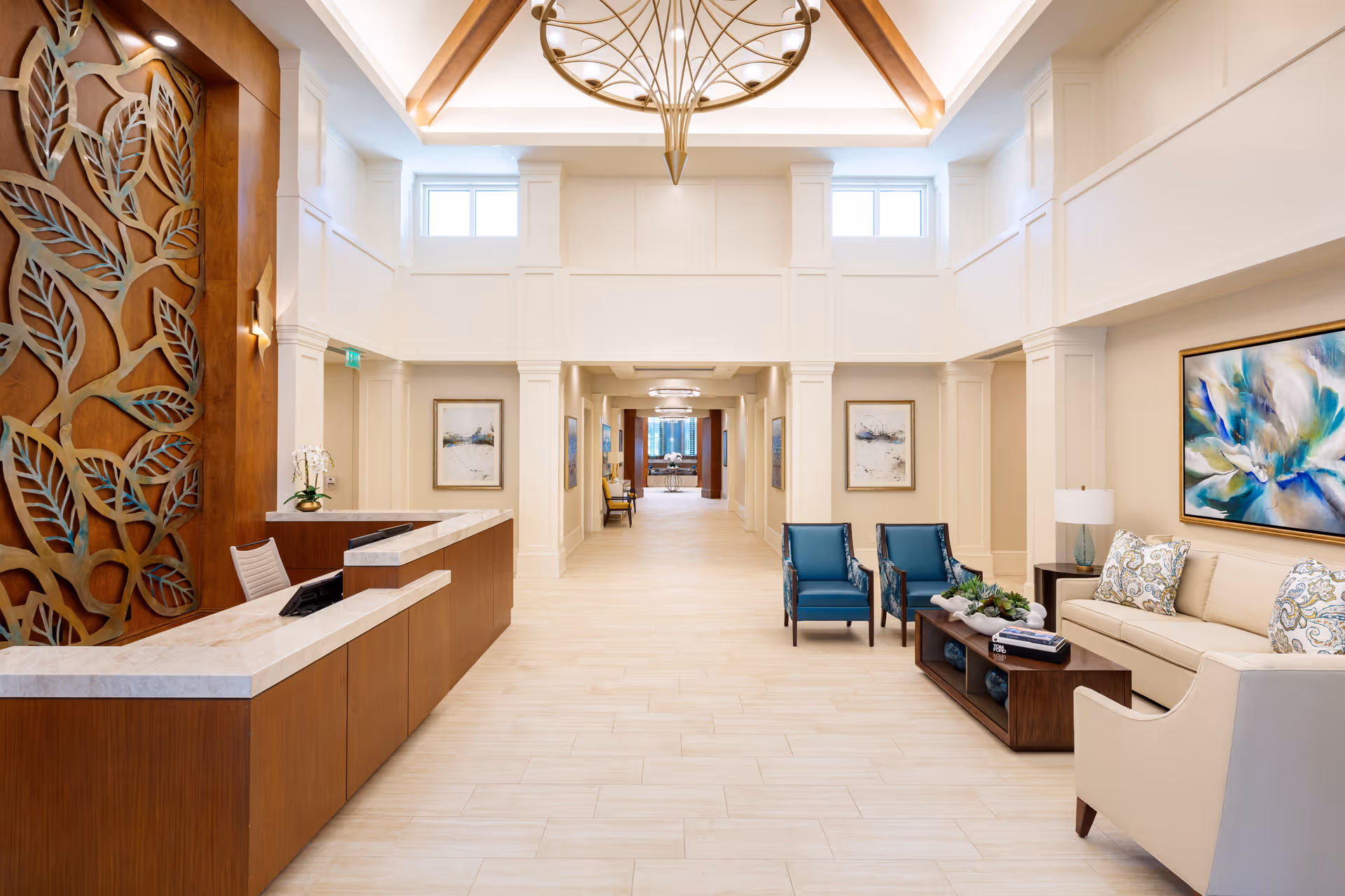 Bright and spacious reception area with a wooden front desk on the left featuring a decorative leaf design panel behind it. The room has high ceilings with large windows near the ceiling, a chandelier, and light-colored tiled floors. On the right, there is a seating area with a beige sofa, two blue armchairs, a beige armchair, a coffee table with decorative items, and colorful artwork on the walls.