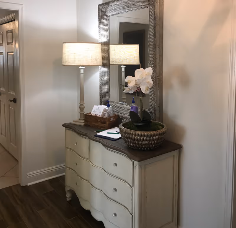A hallway corner featuring a cream-colored dresser with a dark wooden top. On the dresser, there is a decorative lamp with a beige shade, a large rectangular mirror with a textured silver frame, a potted white orchid, a wooden tray holding hand sanitizer and a small card, and a clipboard with a pen. The floor is wooden, and a door is visible on the left side.