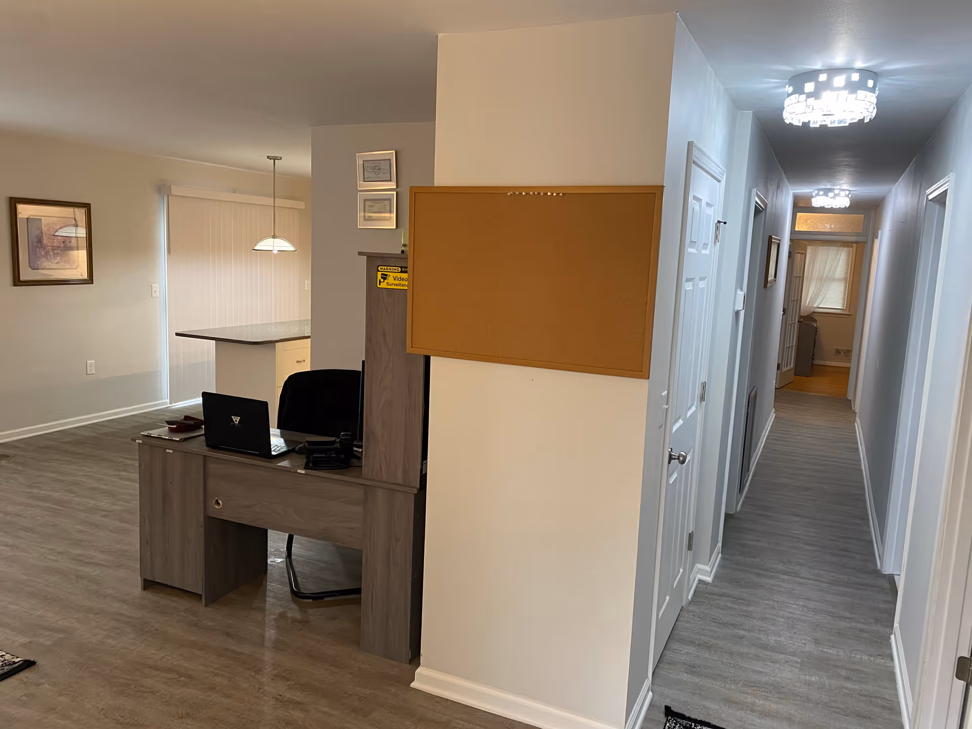 Interior view of a senior living facility showing a reception desk with a laptop and chair, a cork bulletin board on a wall, a hallway with multiple doors and modern ceiling lights, and a dining area with a hanging light fixture and framed artwork on the wall.