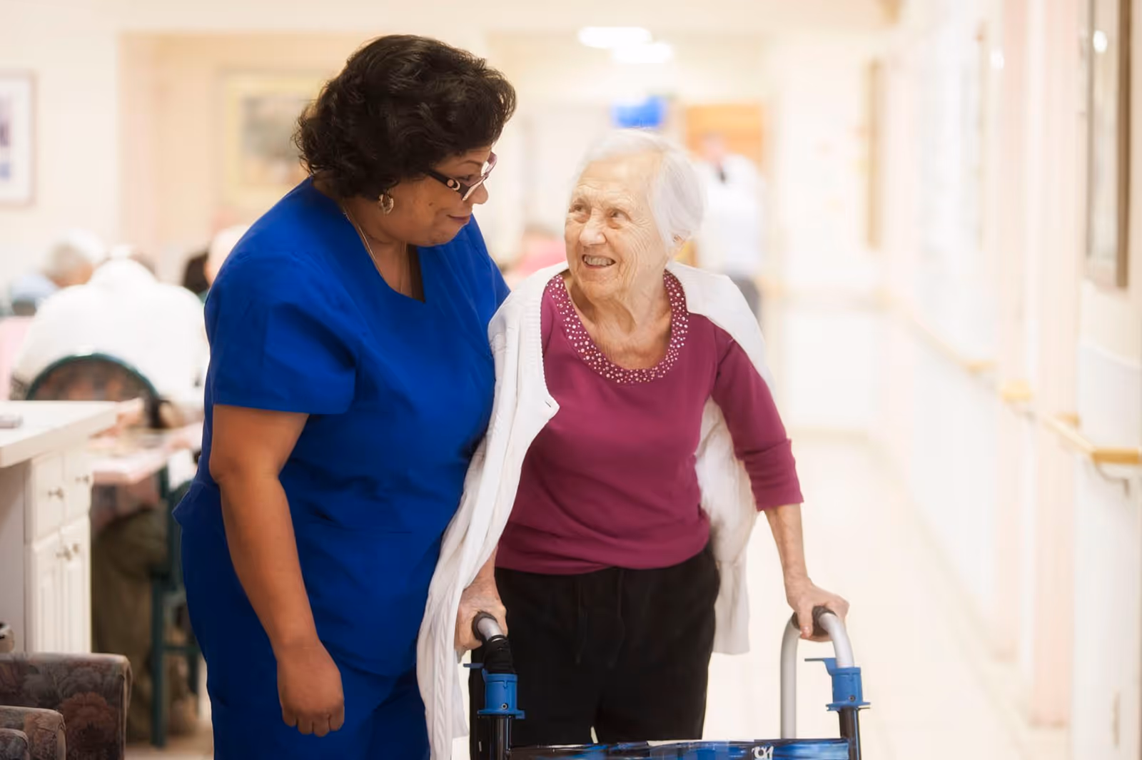 A caregiver in blue scrubs is assisting an elderly woman using a walker in a well-lit hallway of an assisted living facility. The elderly woman is smiling and wearing a purple top with a white cardigan draped over her shoulders.
