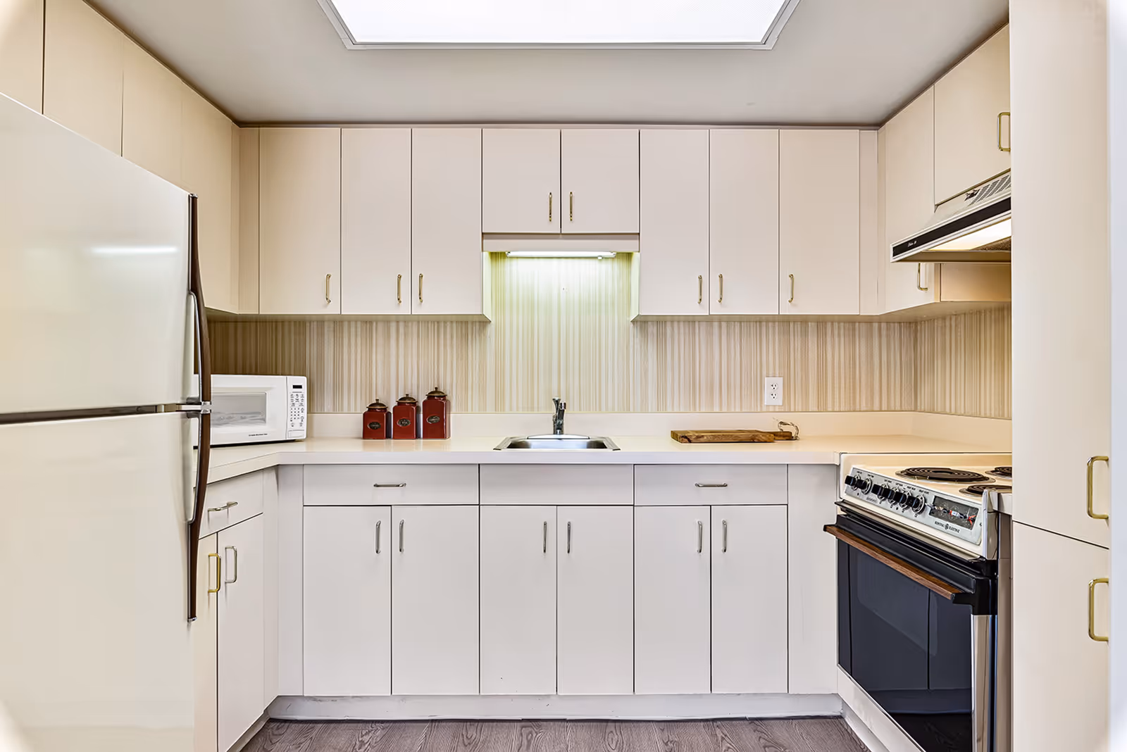 A clean, well-lit kitchen with white cabinets and drawers, a white refrigerator on the left, a microwave on the countertop, a stainless steel sink in the center, and an electric stove with an oven on the right. The backsplash has a vertical striped pattern, and the floor is a dark wood finish.