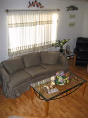 A cozy living room with a beige patterned sofa, a glass coffee table with a floral centerpiece and magazines, a black recliner chair, a potted plant on a small side table, and a window with sheer curtains letting in natural light. The walls have decorative signs and a small floral arrangement above the window.