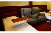 A nursing center room featuring a hospital bed with an overbed table, two cushioned recliner chairs, and a bedside table with a lamp. The walls are painted yellow and brown, and the floor is carpeted in red.