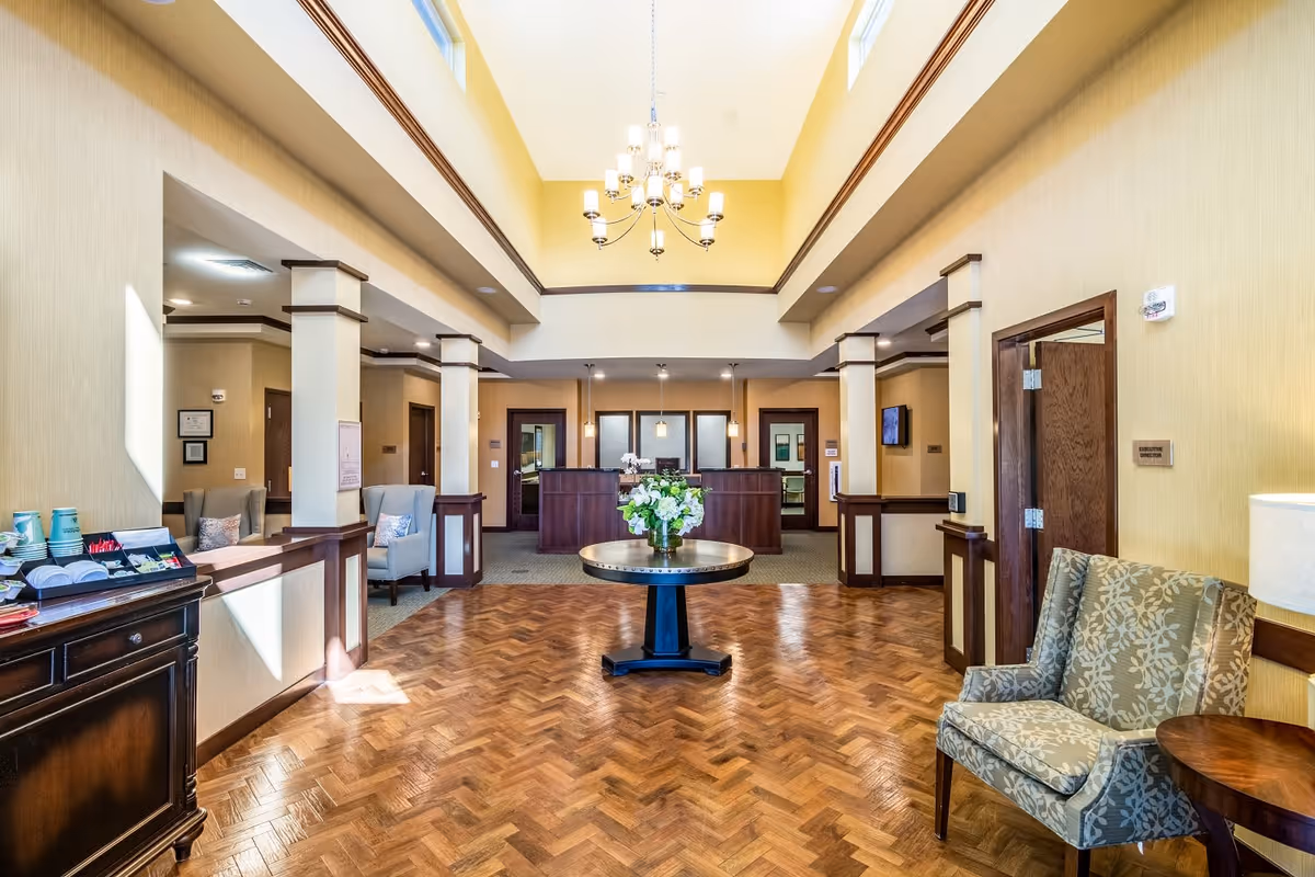 A spacious and well-lit reception area with a high ceiling and chandelier. The room features a round table with a floral arrangement in the center, wood parquet flooring, beige walls with dark wood trim, and several seating areas with upholstered chairs. A reception desk is visible in the background along with doorways leading to other rooms.