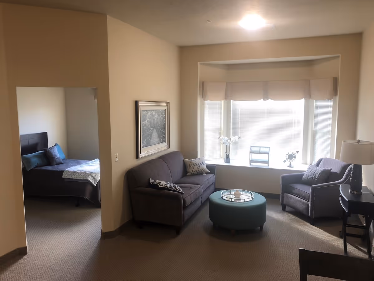 A cozy living area with a gray sofa and armchair, a round teal ottoman with a tray on top, a side table with a lamp, and a large window with blinds and a valance. To the left, there is an open doorway leading to a bedroom with a bed made with dark bedding and pillows. The walls are beige and the carpet is light brown.