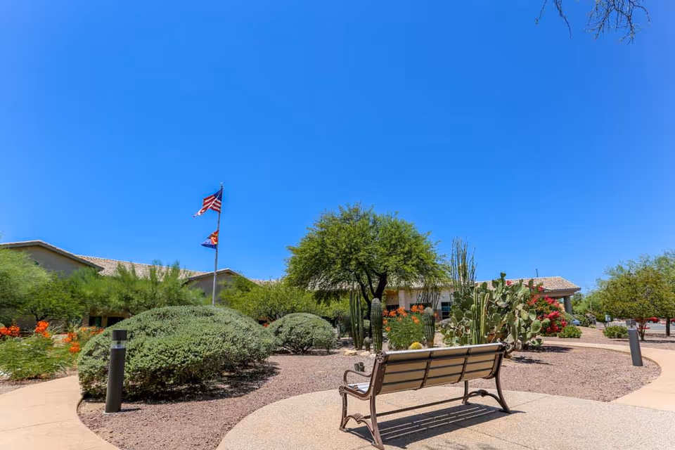Sunny outdoor courtyard with a bench, desert landscaping and cacti, flagpoles, and a single-story building in the background.