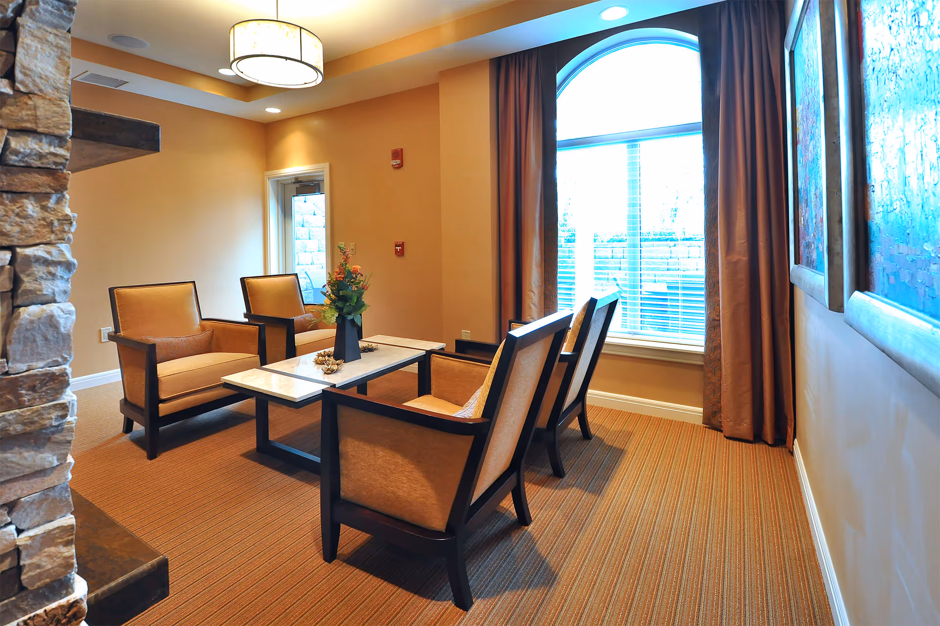 A cozy seating area in a senior living facility with four cushioned armchairs arranged around a rectangular coffee table with a flower vase centerpiece. The room has warm beige walls, a large arched window with brown curtains, a stone fireplace on the left, and framed artwork on the right wall. A modern ceiling light fixture hangs above.