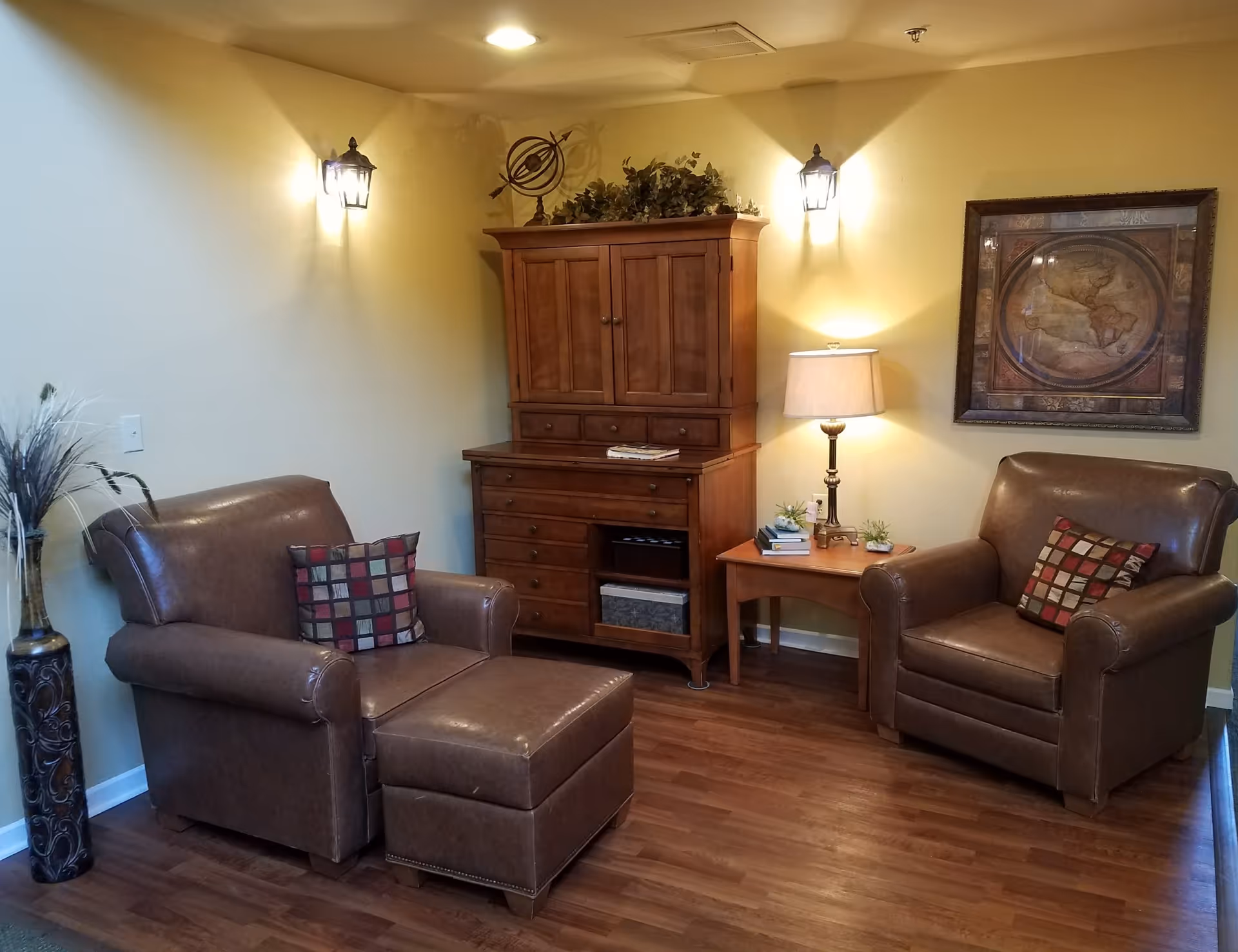 A cozy living room corner with two brown leather armchairs, one with a matching ottoman, each with a decorative square-patterned pillow. Between the chairs is a wooden side table with a lamp, books, and small plants. Behind the table is a wooden cabinet topped with greenery and a decorative armillary sphere. A framed vintage map hangs on the wall, and two wall-mounted lantern-style lights illuminate the space. The floor is wood, and a tall decorative vase with dried plants stands to the left.