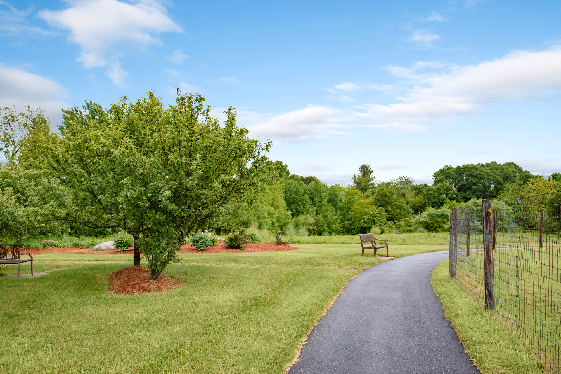A paved walking path curves through a green outdoor area with grass, trees, and benches. A wire fence runs along the right side of the path, and the sky is partly cloudy.