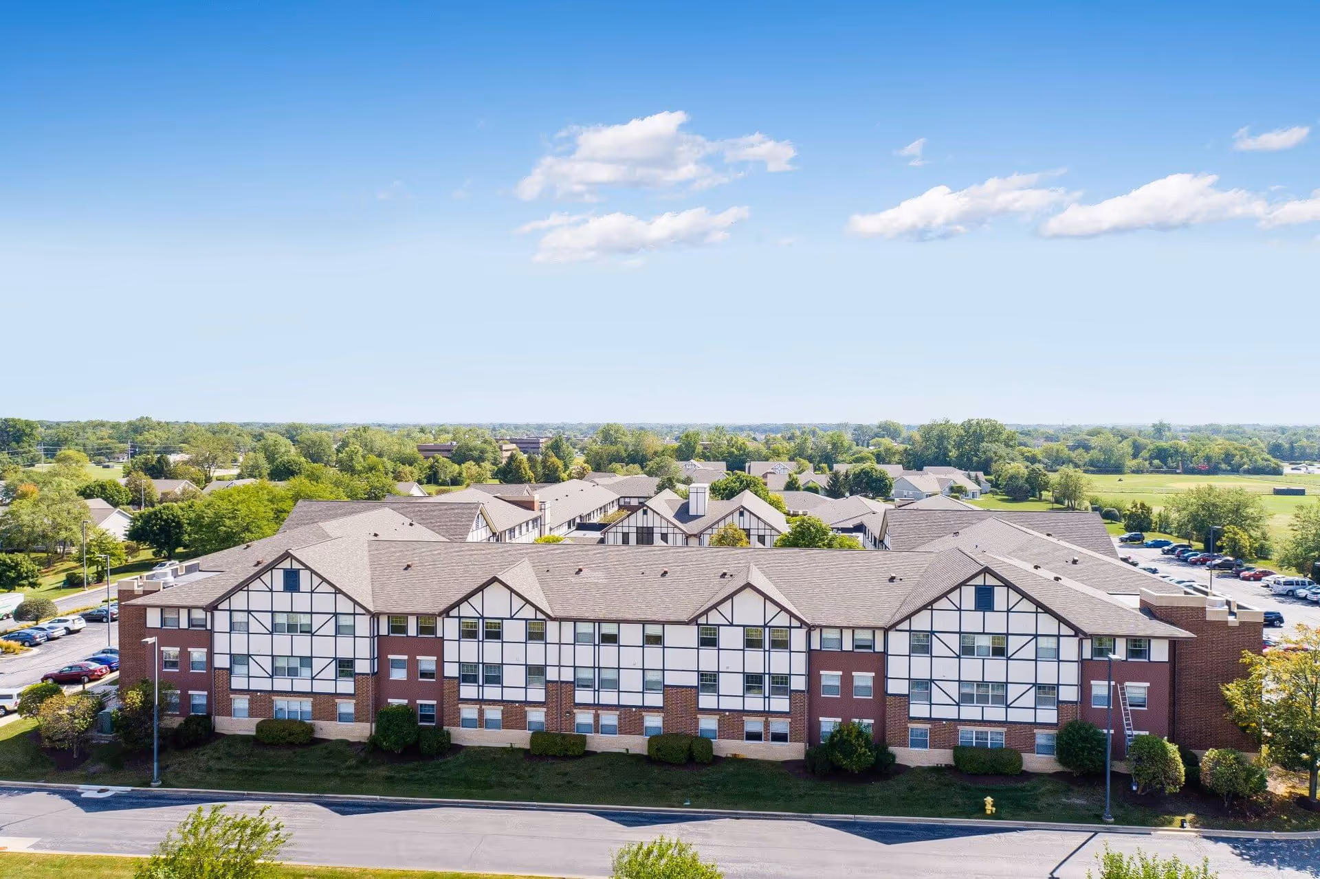 Aerial view of a large senior living facility building with Tudor-style architecture, featuring white walls with dark wooden beams and a brown roof. The building is surrounded by greenery, trees, and a parking lot with cars. The sky is clear with a few clouds.