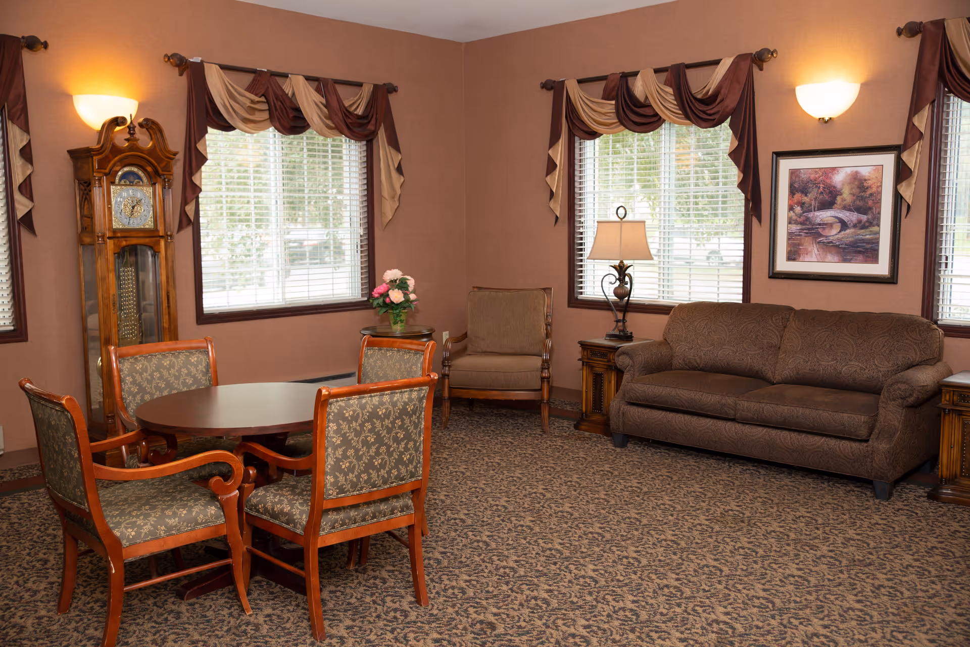 A furnished common living room with a sofa, armchairs, a round table with chairs, a grandfather clock, lamps, and windows with draped curtains.