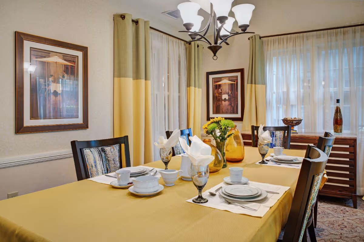 A dining room with a yellow tablecloth-covered table set for a meal, chairs, vases of flowers, framed artwork, and a chandelier.