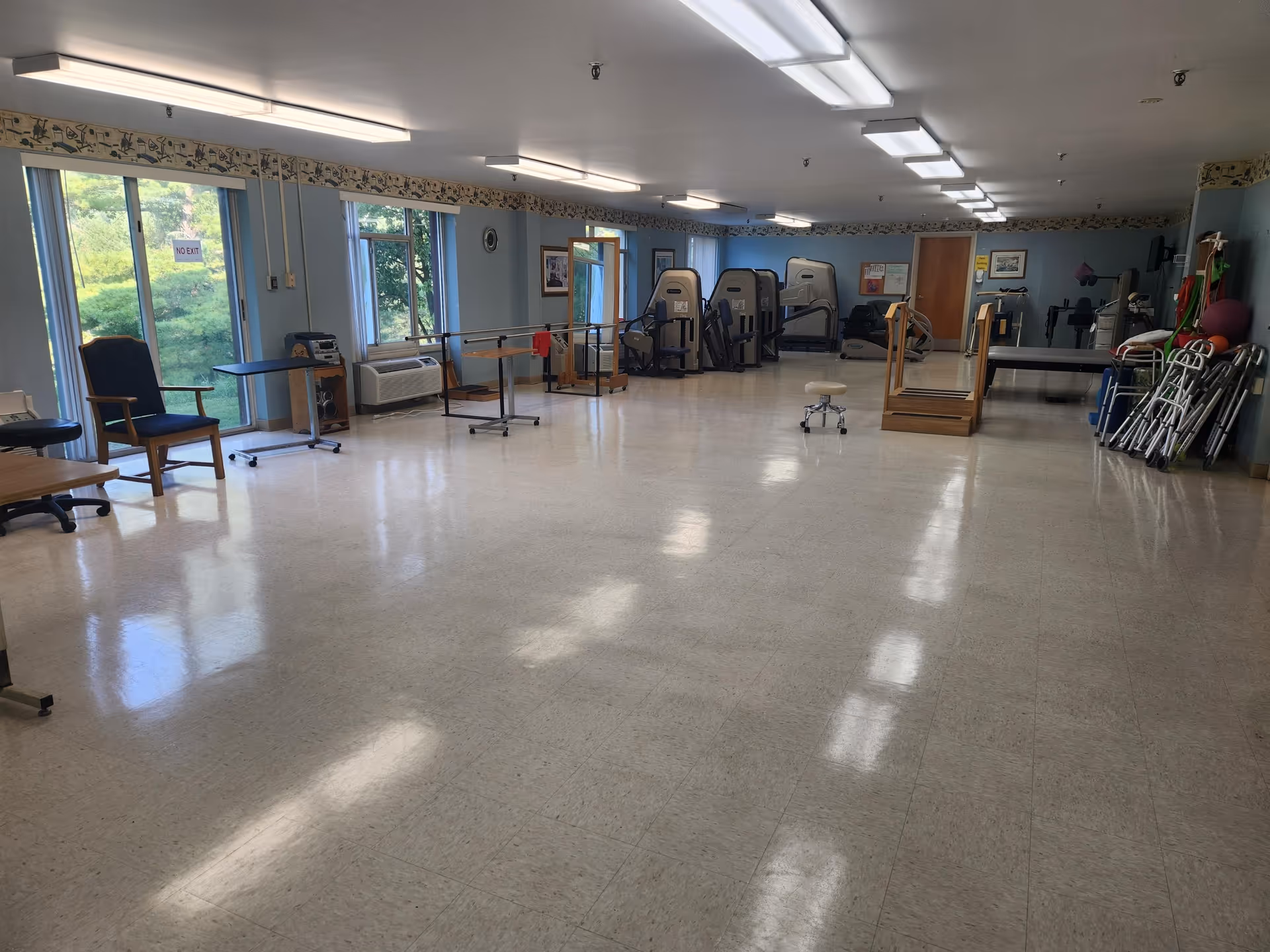 A spacious rehabilitation or physical therapy room with exercise equipment including treadmills, parallel bars, and various mobility aids such as walkers and canes. The room has large windows letting in natural light, light blue walls, and a tiled floor.