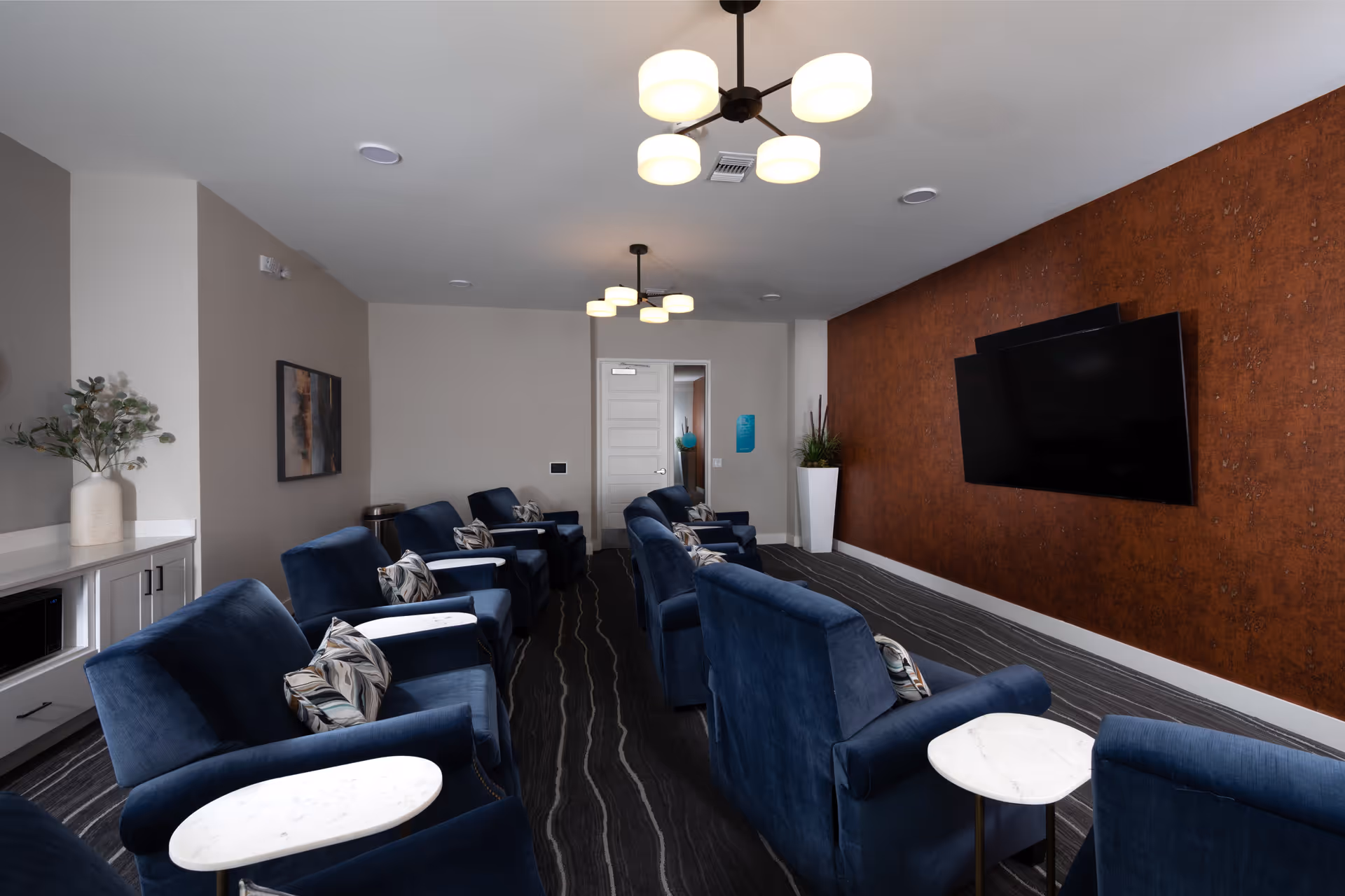 A modern lounge room with two rows of blue upholstered armchairs, each with a small white marble side table. The room features a large flat-screen TV mounted on a rust-colored accent wall, a white door at the back, and two ceiling light fixtures with round shades. There is a white cabinet with a microwave and a vase with greenery on the left side.