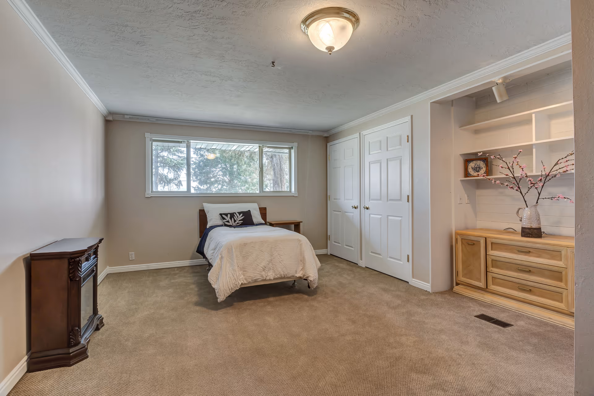 A simple bedroom with a single bed covered in white bedding and a decorative pillow, a wooden nightstand, a built-in wooden cabinet with shelves holding a vase with pink flowers and a decorative plate, beige carpet, a window with a view of trees, and a ceiling light fixture.
