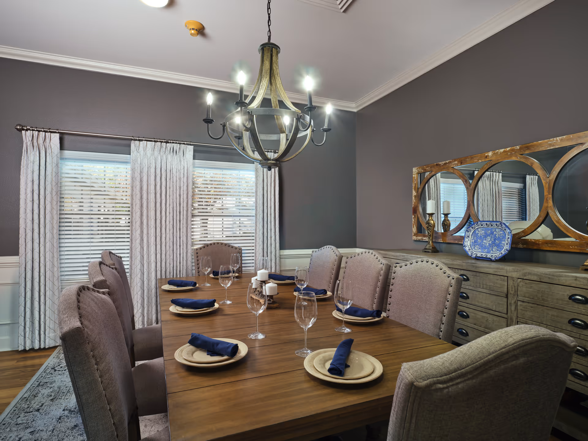 A dining room with a wooden table set for six, featuring beige upholstered chairs, plates with navy blue napkins, and wine glasses. The room has gray walls, white curtains on the windows, a decorative chandelier hanging from the ceiling, and a wooden sideboard with a large mirror and decorative items.