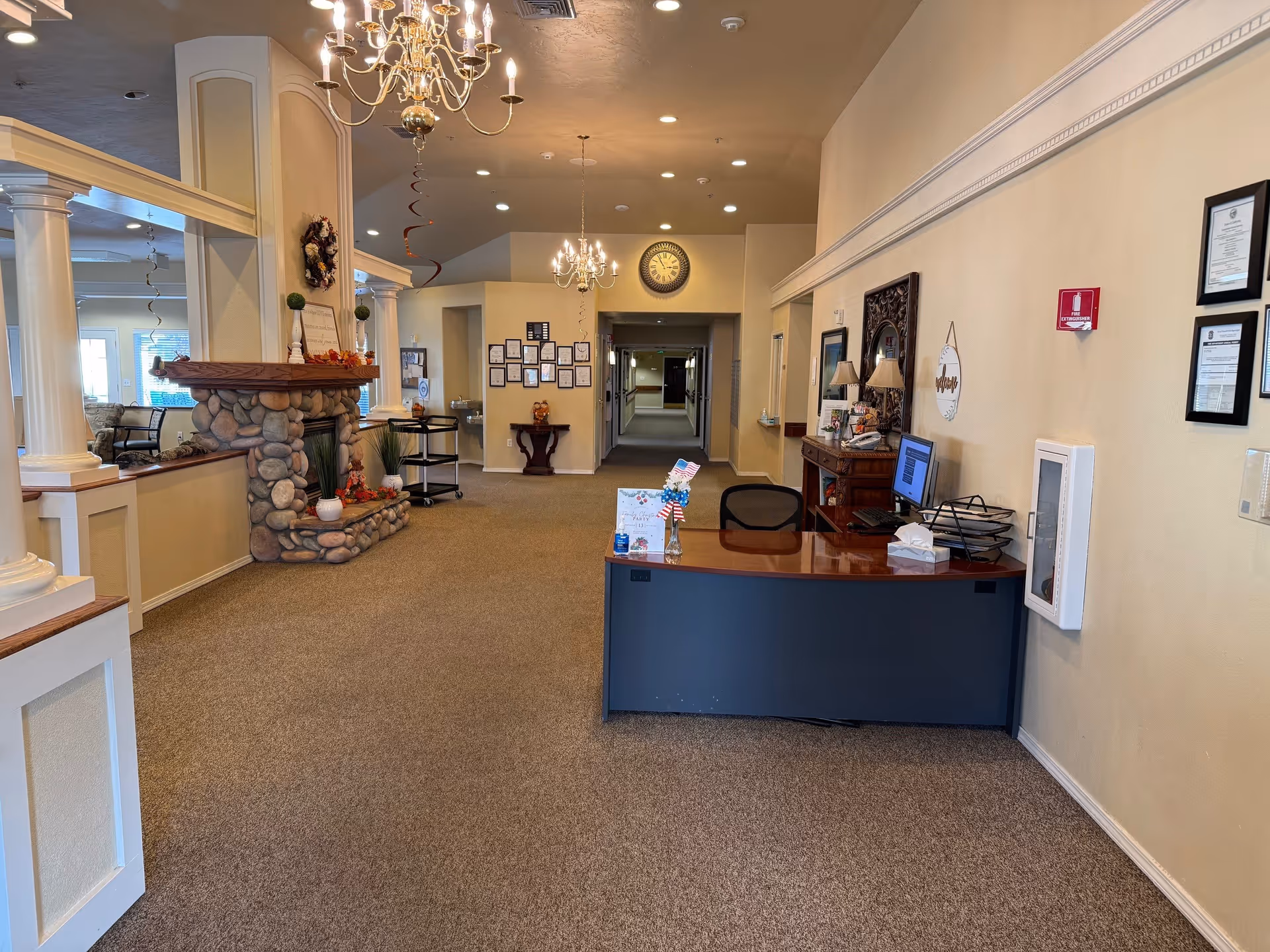 Interior view of a senior living facility reception area with a desk, computer, and chair on the right side. The room features beige walls, carpeted floor, chandeliers hanging from the ceiling, a stone fireplace decorated with autumn-themed items, and a hallway leading further into the building. Various framed certificates and decorations are mounted on the walls.